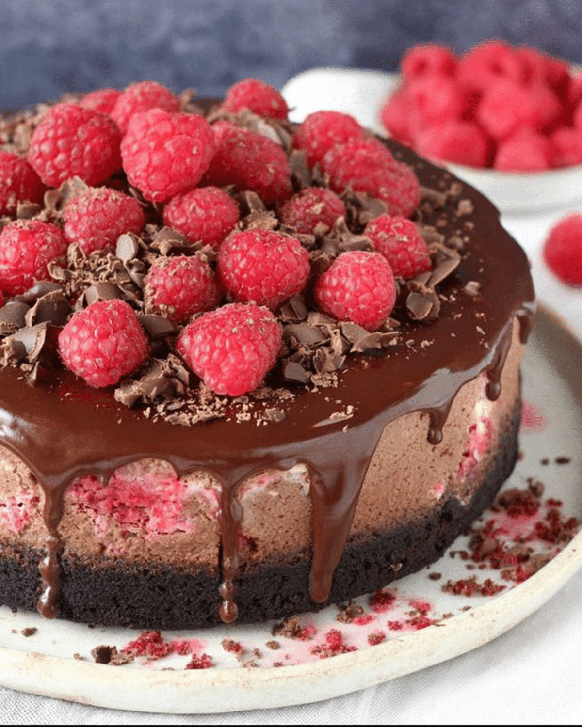 Chocolate raspberry cheesecake topped with fresh raspberries, chocolate shavings, and dripping chocolate ganache on a cookie crust.