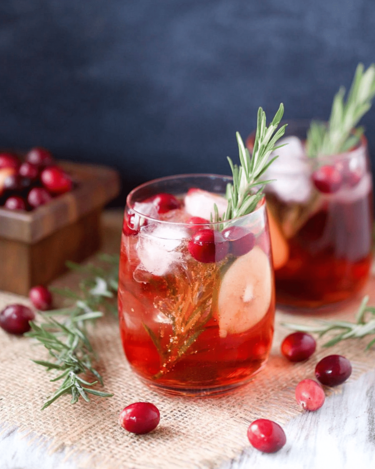 Christmas Apple Cranberry Spritzer in a glass with ice, fresh cranberries, apple slices, and rosemary garnish.