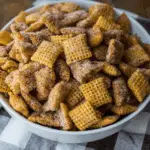 A bowl filled with Churro Chex Mix, coated in cinnamon sugar, sitting on a checkered cloth.