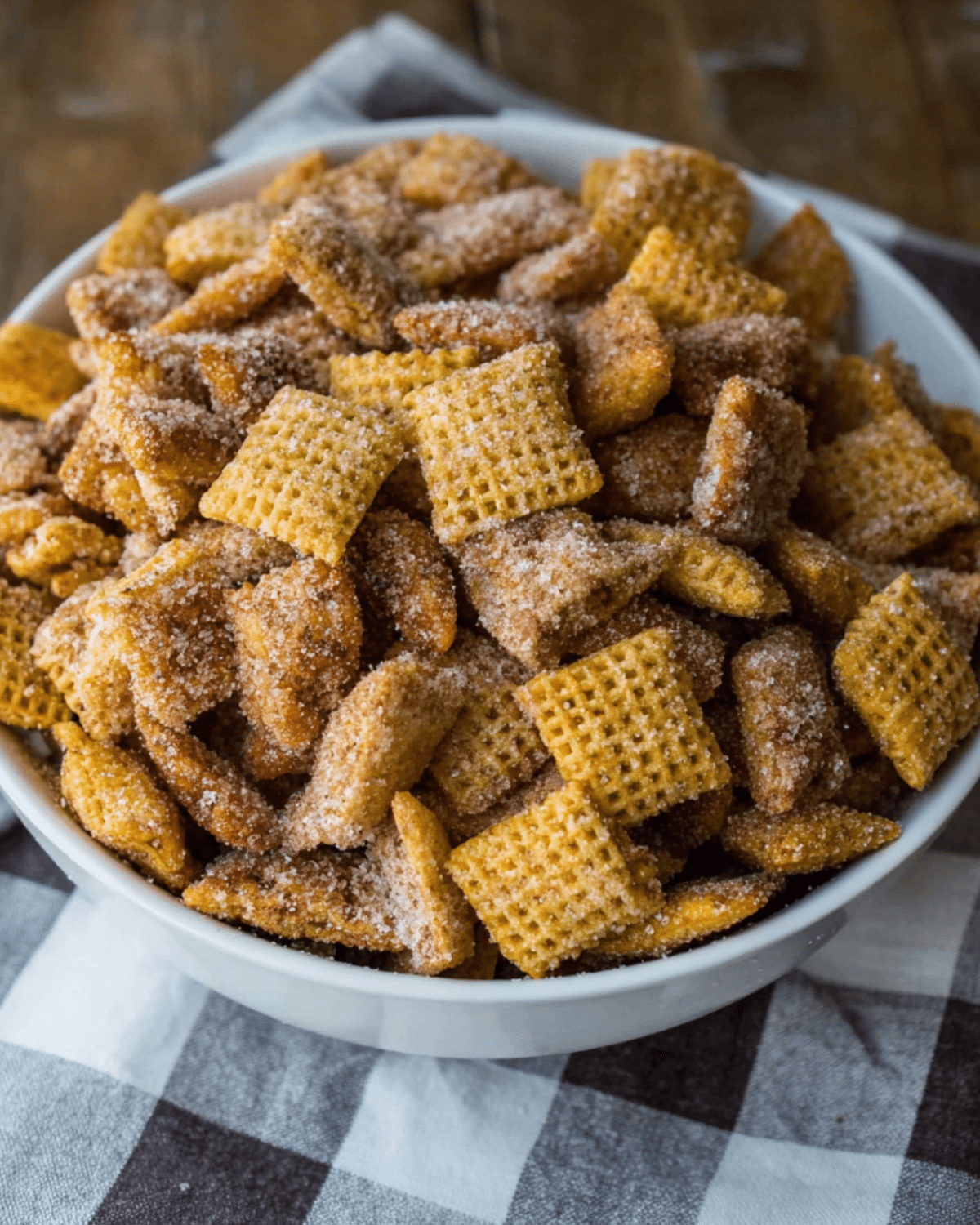 A bowl filled with Churro Chex Mix, coated in cinnamon sugar, sitting on a checkered cloth.