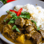 Plate of coconut beef curry with white rice, garnished with cilantro and sliced red chili peppers.