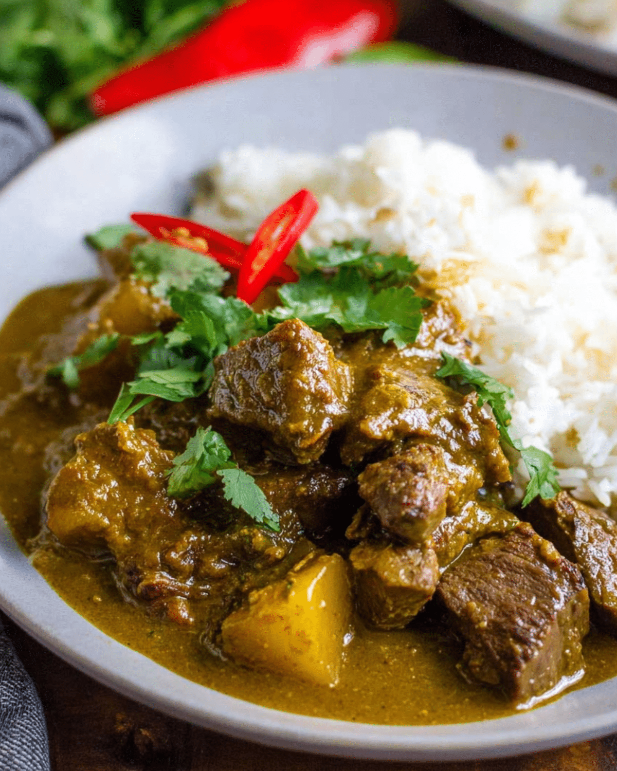Plate of coconut beef curry with white rice, garnished with cilantro and sliced red chili peppers.