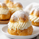 Golden cream puff filled with piped whipped cream and dusted with powdered sugar on a white plate.