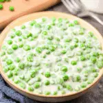 Bowl of creamed peas in a rich, white sauce placed on a blue cloth napkin with a fork and scattered peas in the background.