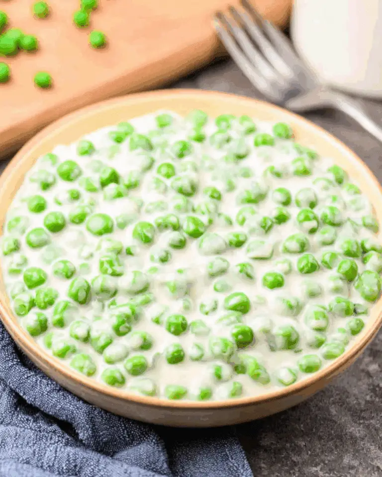 Bowl of creamed peas in a rich, white sauce placed on a blue cloth napkin with a fork and scattered peas in the background.