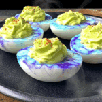 Creepy Deviled Eggs for Halloween with blue and purple marbled egg whites and green filling, topped with red seasoning on a dark plate.