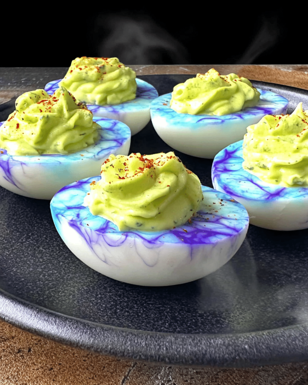 Creepy Deviled Eggs for Halloween with blue and purple marbled egg whites and green filling, topped with red seasoning on a dark plate.