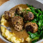 A bowl of easy meatballs and gravy served with mashed potatoes and sautéed greens.