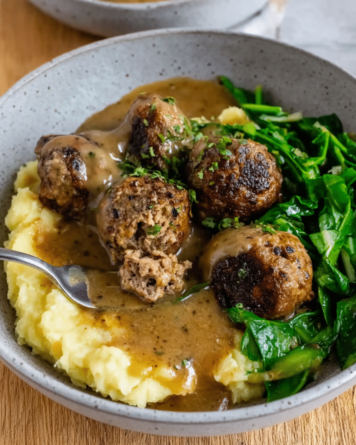 A bowl of easy meatballs and gravy served with mashed potatoes and sautéed greens.
