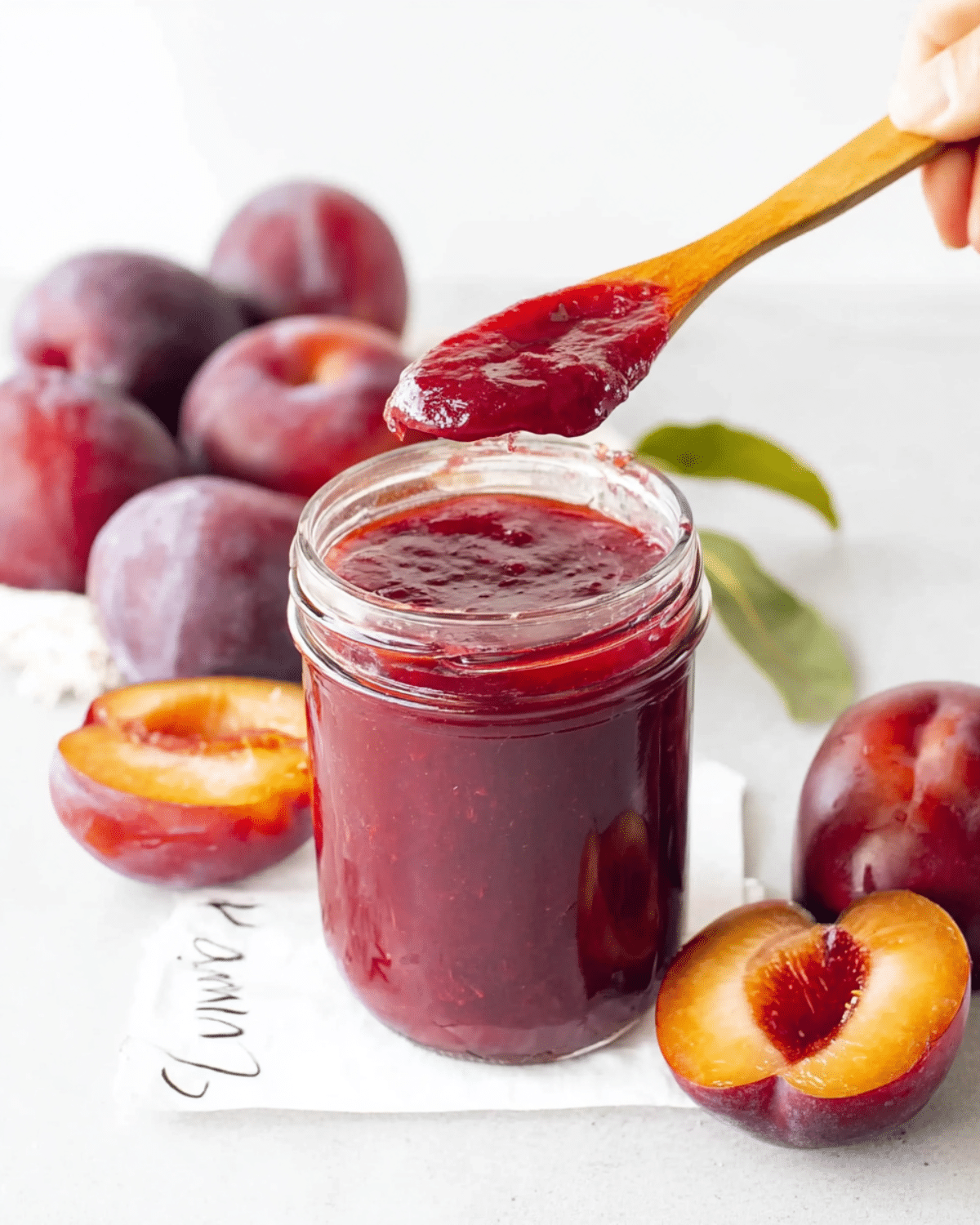 A jar of homemade plum jam with a spoon lifting a thick, glossy scoop, surrounded by fresh whole and halved plums.