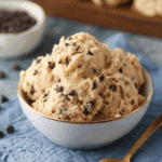 A bowl filled with scoops of edible cookie dough studded with mini chocolate chips, next to a gold spoon.