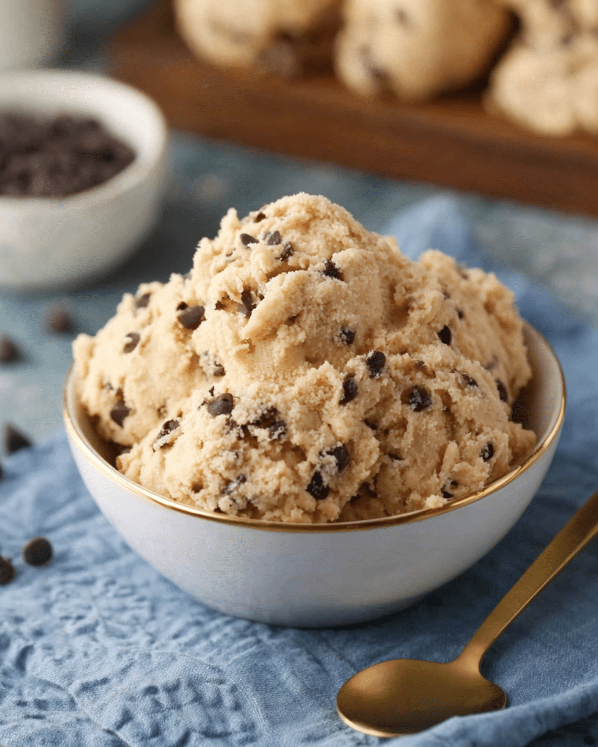 A bowl filled with scoops of edible cookie dough studded with mini chocolate chips, next to a gold spoon.