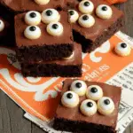 Eyeball Brownies topped with candy eyeballs, stacked on festive Halloween-themed paper with a small pumpkin decoration.