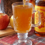 Glass mug of Fireball apple cider being poured, with fresh apples and a Fireball whiskey bottle in the background.