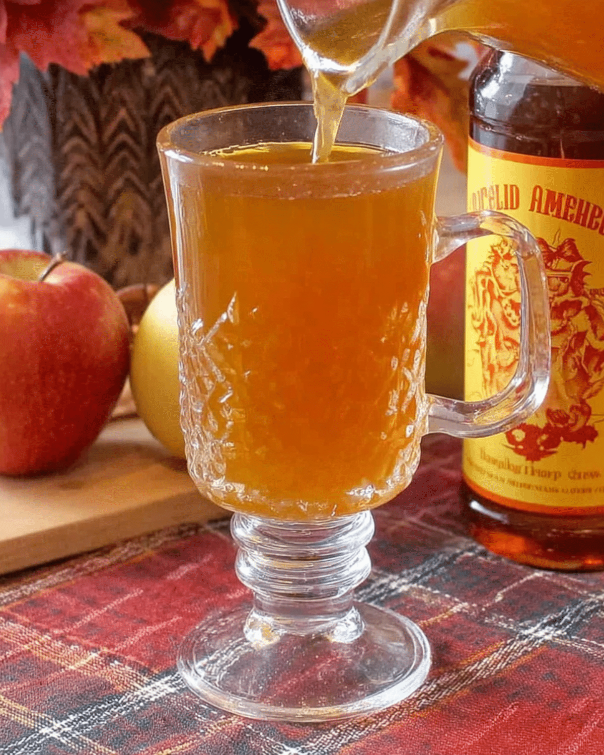 Glass mug of Fireball apple cider being poured, with fresh apples and a Fireball whiskey bottle in the background.