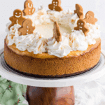 Gingerbread Cheesecake Cake topped with whipped cream swirls and decorated with mini gingerbread cookies on a cake stand.