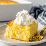 Moist slice of Greek yogurt cake topped with whipped cream on a ceramic plate with a gold fork.