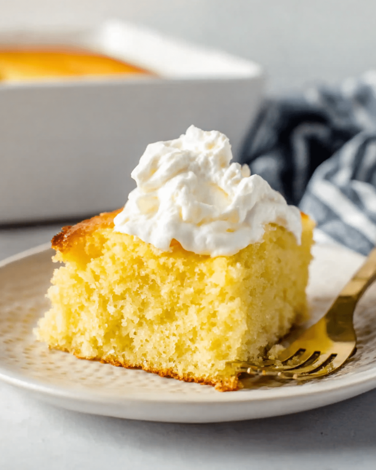 Moist slice of Greek yogurt cake topped with whipped cream on a ceramic plate with a gold fork.