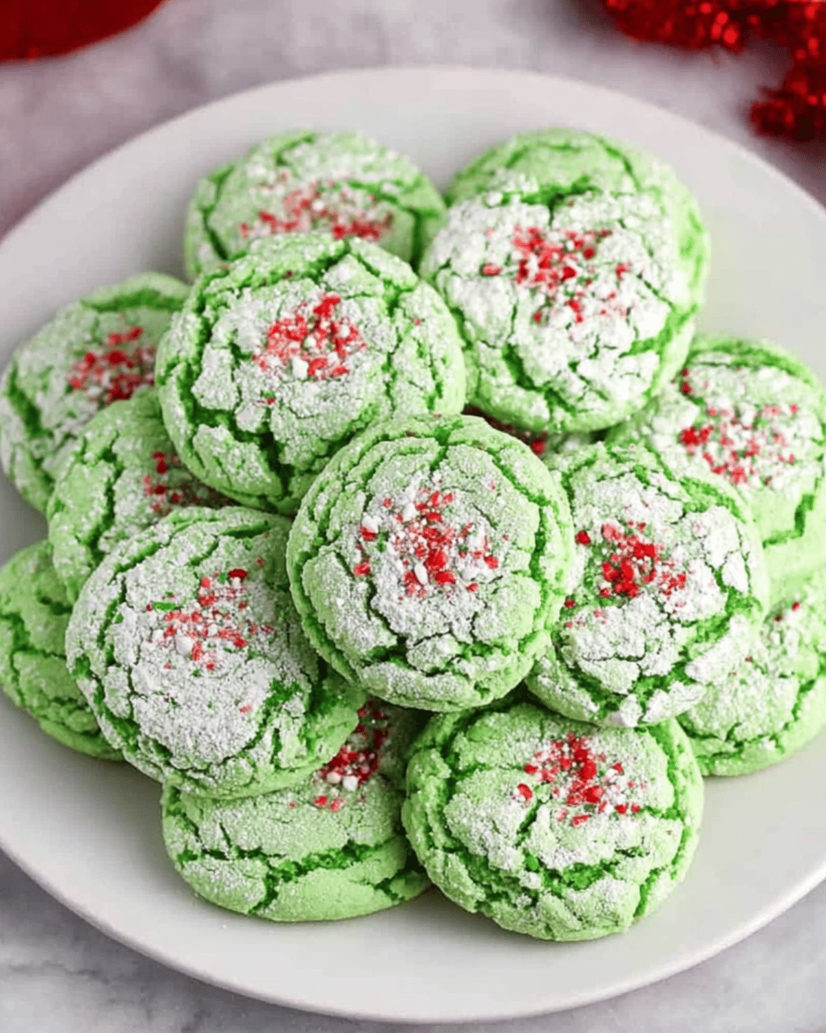 Plate of bright green Grinch Cookie Magic cookies dusted with powdered sugar and topped with red and white sprinkles.