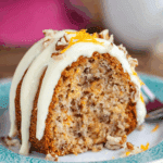 Slice of hummingbird bundt cake topped with cream cheese frosting, chopped pecans, and orange zest on a decorative plate.
