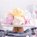 A colorful Jello Divinity Recipe dessert displayed on a white cake stand, featuring fluffy pastel swirls in pink, yellow, blue, and white.