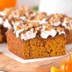 A slice of keto pumpkin cake topped with white frosting, chopped nuts, and cinnamon on a wooden board.