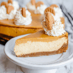 Slice of layered pumpkin cheesecake topped with whipped cream and a gingersnap cookie, with the whole cheesecake in the background.