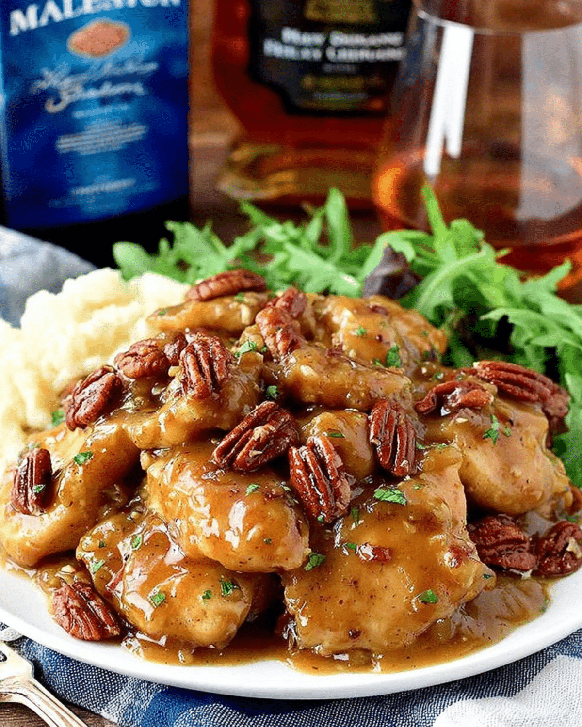 A plate of maple bourbon pecan chicken covered in a rich glaze, served with mashed potatoes and greens.