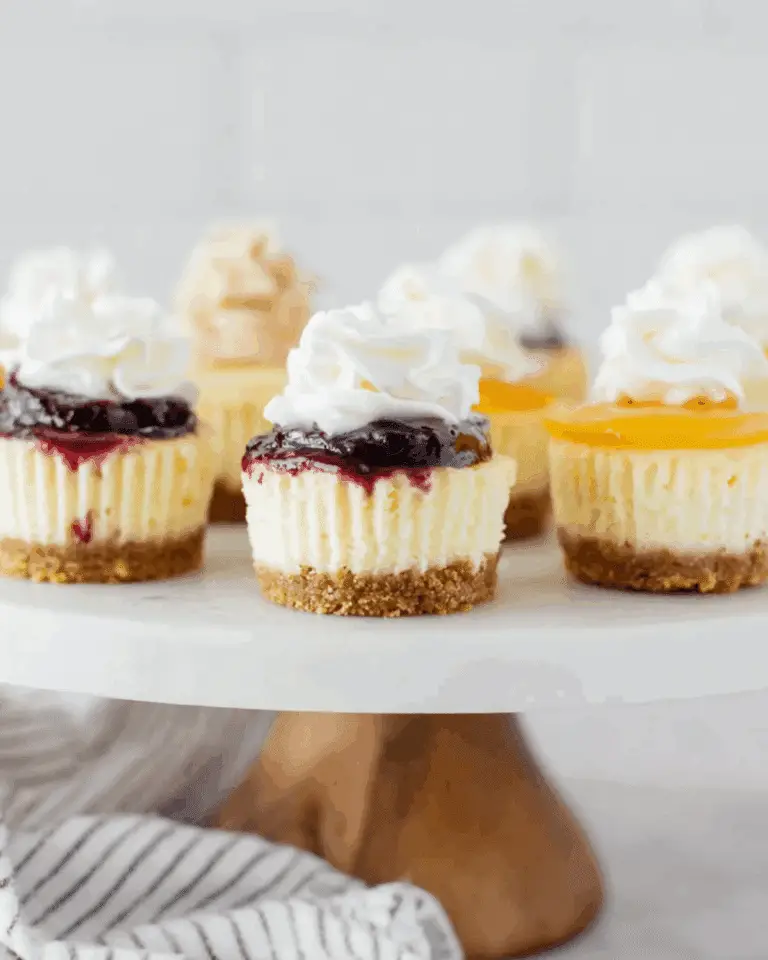 Assorted mini cheesecakes topped with whipped cream and various fruit toppings like blueberry and peach on a cake stand.
