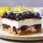 No-bake lemon blueberry delight dessert with a graham cracker crust, creamy lemon filling, blueberry layers, and fresh blueberries with lemon zest on top.