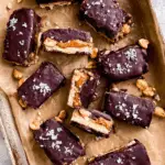 Homemade No bake Snickers Bars topped with flaky sea salt, filled with caramel, peanuts, and chocolate on a parchment-lined tray.