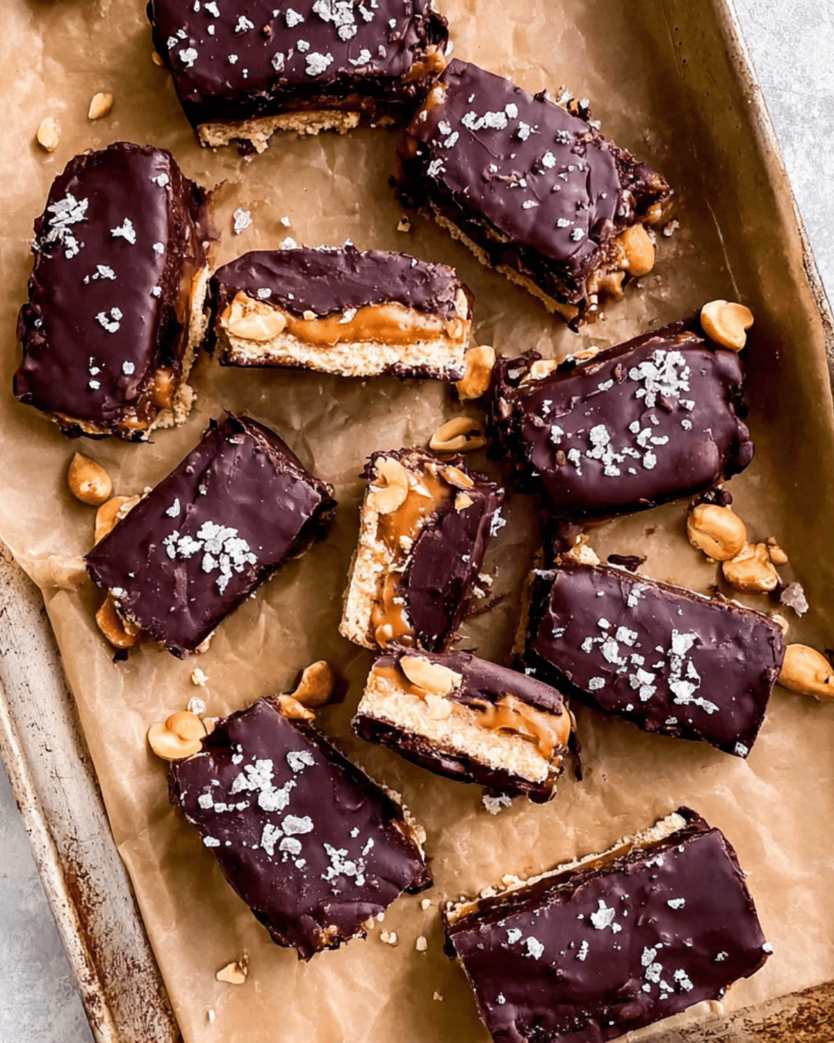 Homemade No bake Snickers Bars topped with flaky sea salt, filled with caramel, peanuts, and chocolate on a parchment-lined tray.