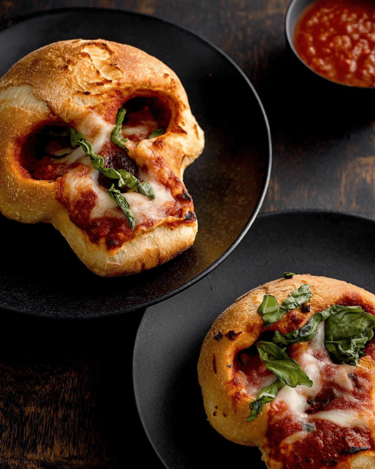 Pizza Skulls made with golden baked dough shaped like skulls, filled with marinara sauce, melted cheese, and fresh basil, served with a side of dipping sauce.