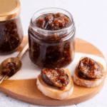 A jar of preserved figs with visible seeds and syrup, accompanied by fig-topped bread slices and a spoonful of the preserve on a wooden board.