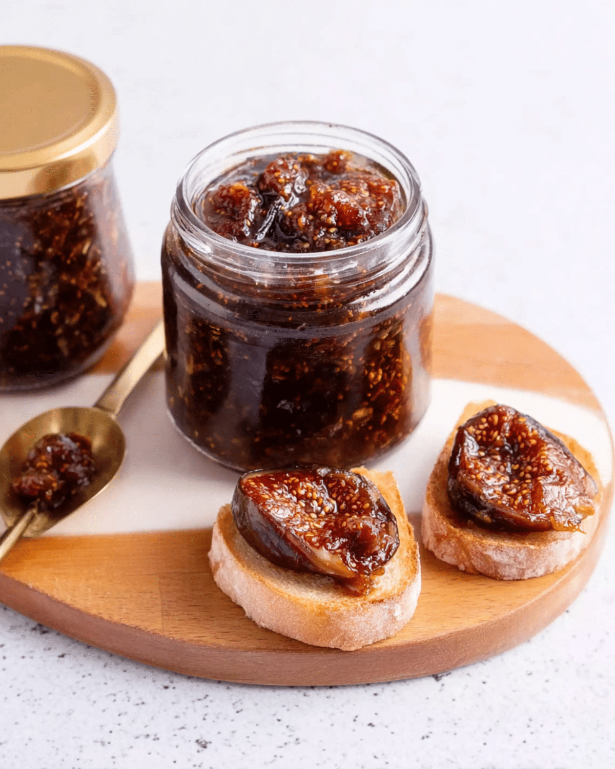 A jar of preserved figs with visible seeds and syrup, accompanied by fig-topped bread slices and a spoonful of the preserve on a wooden board.
