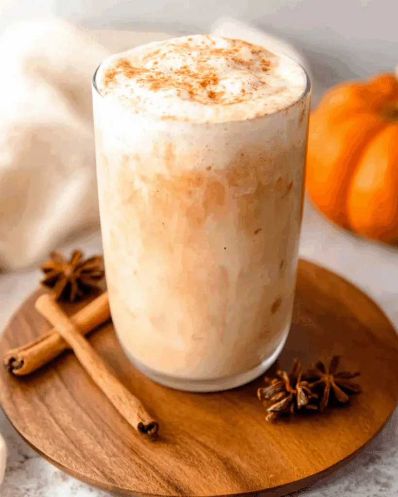 Pumpkin chai iced latte topped with creamy pumpkin foam and sprinkled with cinnamon, served on a wooden coaster with cinnamon sticks and star anise.
