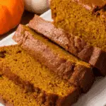 Sliced pumpkin loaf on a white plate, with whole pumpkins in the background.