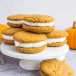 Pumpkin pie cookie sandwiches filled with creamy white frosting, stacked on a white cake stand.