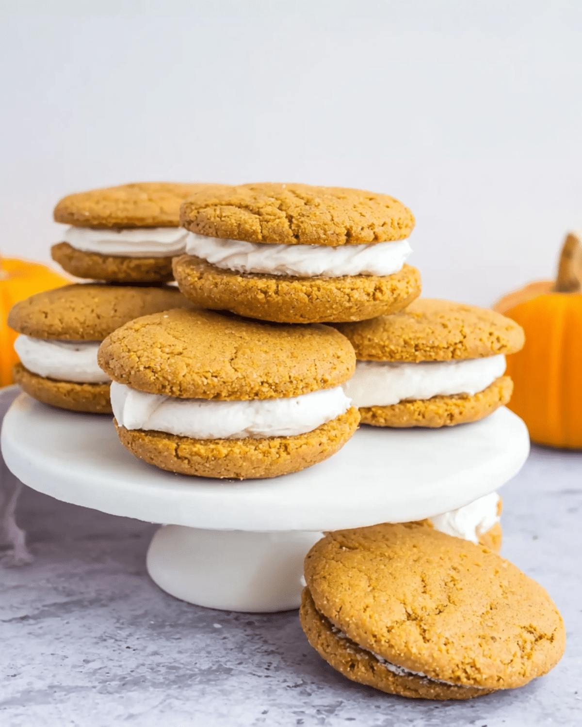 Pumpkin pie cookie sandwiches filled with creamy white frosting, stacked on a white cake stand.