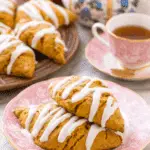 Pumpkin scones topped with white icing drizzle, served on a pink floral plate beside a cup of tea and a teapot.