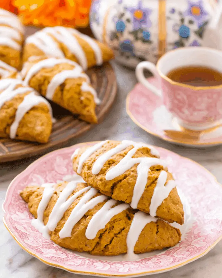 Pumpkin scones topped with white icing drizzle, served on a pink floral plate beside a cup of tea and a teapot.