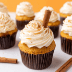 Pumpkin spice cupcake topped with swirled tangy frosting, dusted with cinnamon, and garnished with a cinnamon stick, surrounded by more cupcakes.