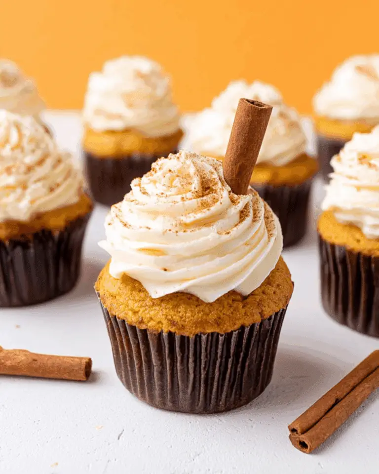 Pumpkin spice cupcake topped with swirled tangy frosting, dusted with cinnamon, and garnished with a cinnamon stick, surrounded by more cupcakes.