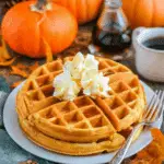 Pumpkin spice waffles topped with whipped cream on a white plate, surrounded by pumpkins, maple syrup, and autumn leaves.