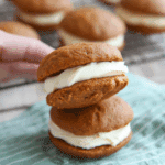 Pumpkin whoopie pies filled with creamy cream cheese frosting, stacked on a striped cloth with more pies cooling on a rack in the background.