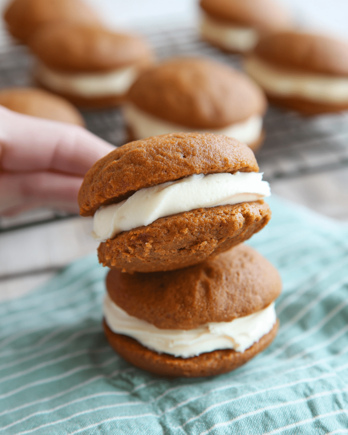 Pumpkin whoopie pies filled with creamy cream cheese frosting, stacked on a striped cloth with more pies cooling on a rack in the background.