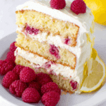 Slice of raspberry cake with lemon buttercream, garnished with fresh raspberries and lemon slices on a white plate.