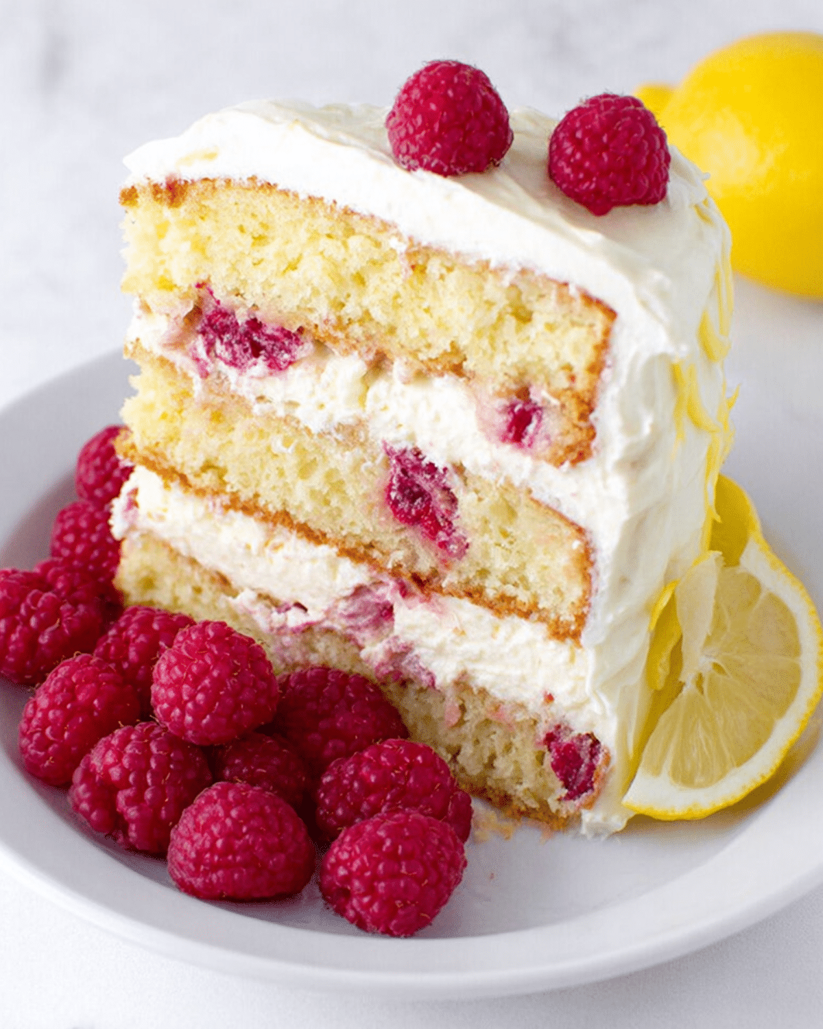 Slice of raspberry cake with lemon buttercream, garnished with fresh raspberries and lemon slices on a white plate.
