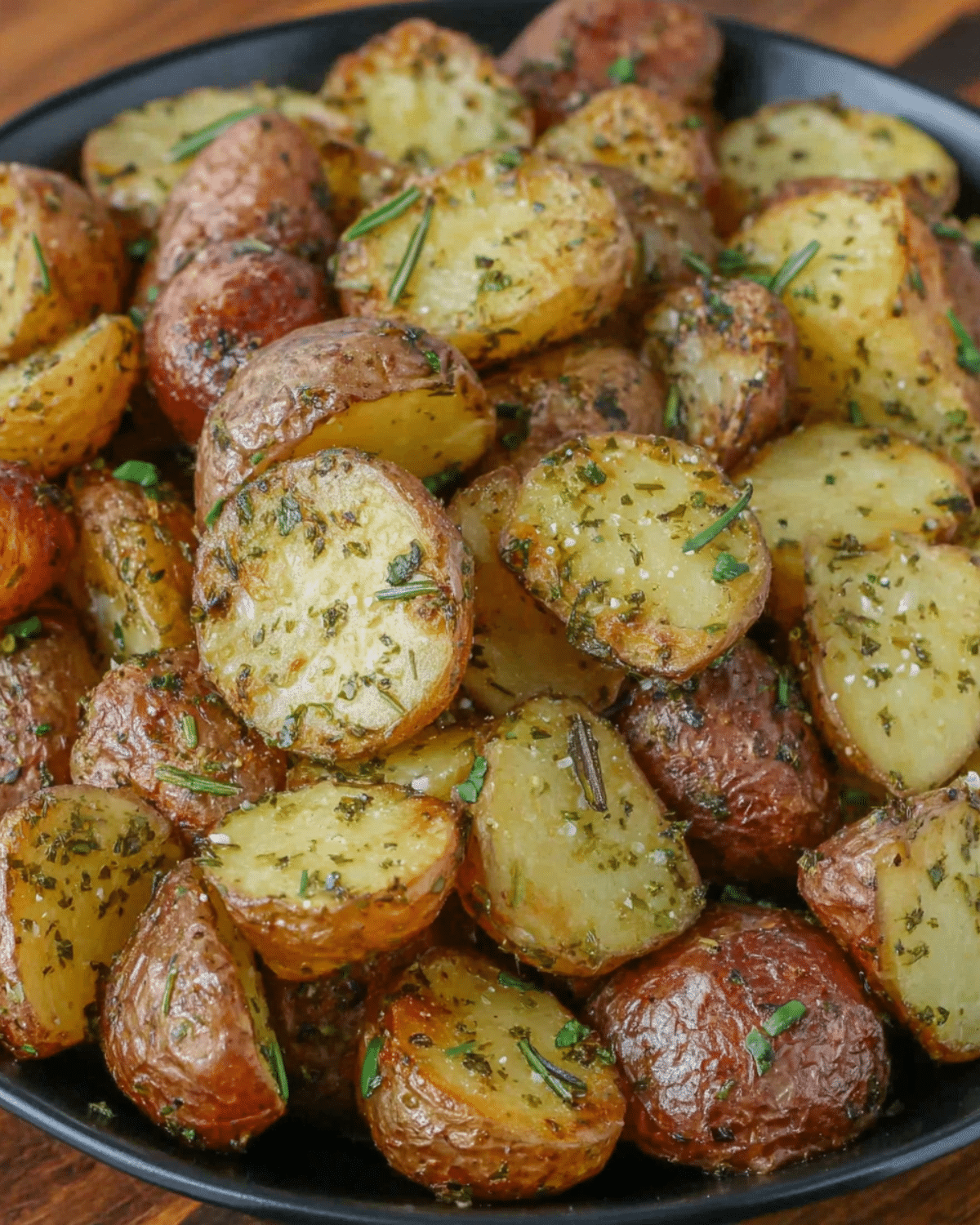 Crispy golden roast potatoes halved and coated with rosemary and herbs, served on a black plate.