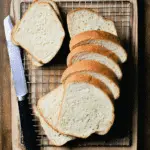 Slices of soft sourdough sandwich bread on a cooling rack with a bread knife on a wooden surface.